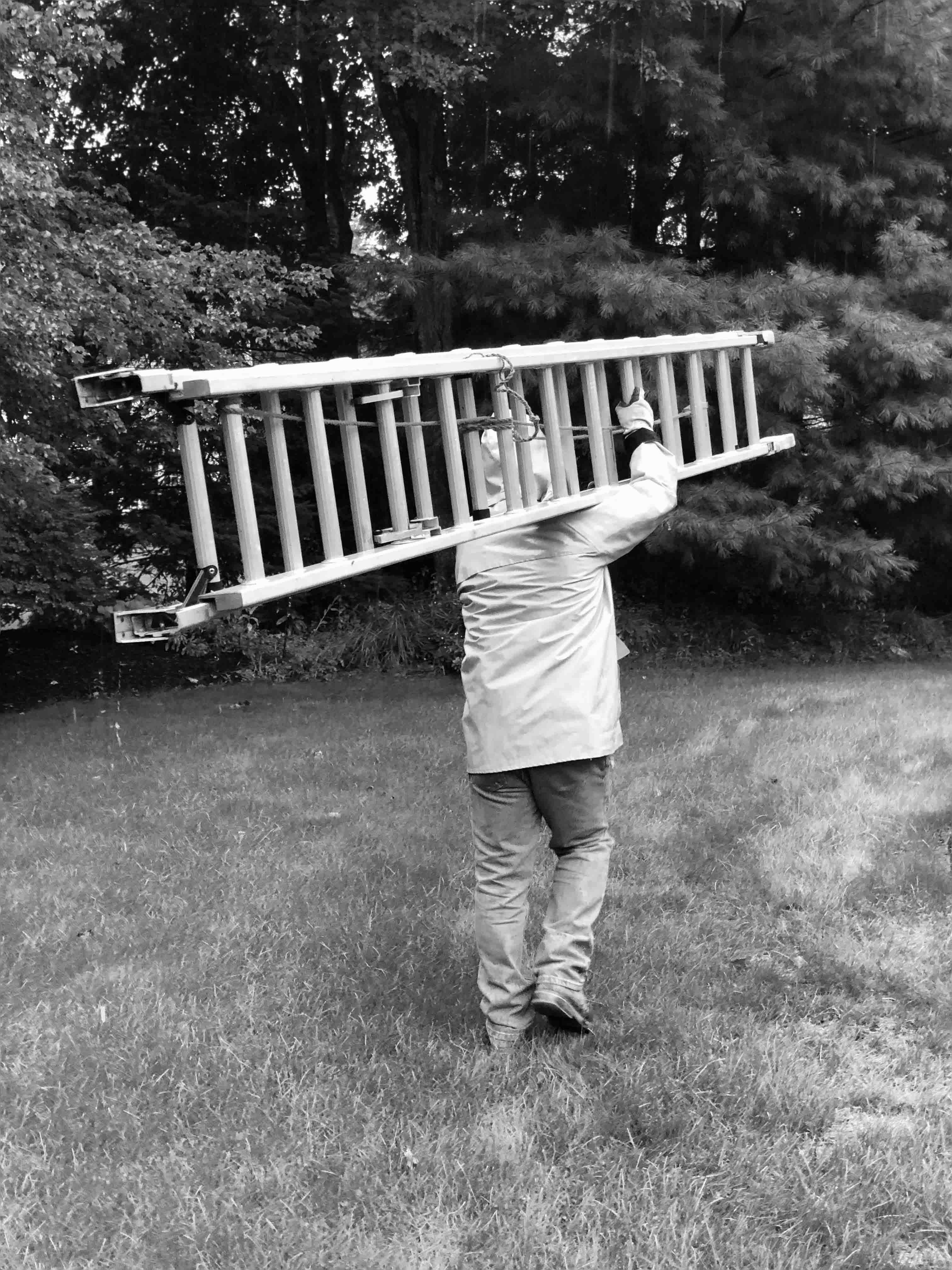 A man in black and white walking with a ladder on his left shoulder